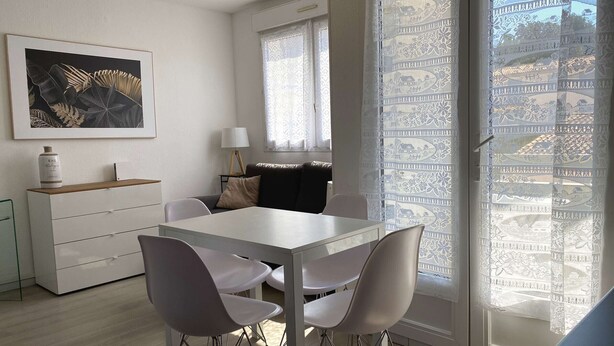 A modern living room with a dining area, featuring a white table, grey chairs, a sofa, and a decorative plant. - Charming T2 Apartment with Private Garage and Balcony in Central Village (Balaruc-les-Bains)