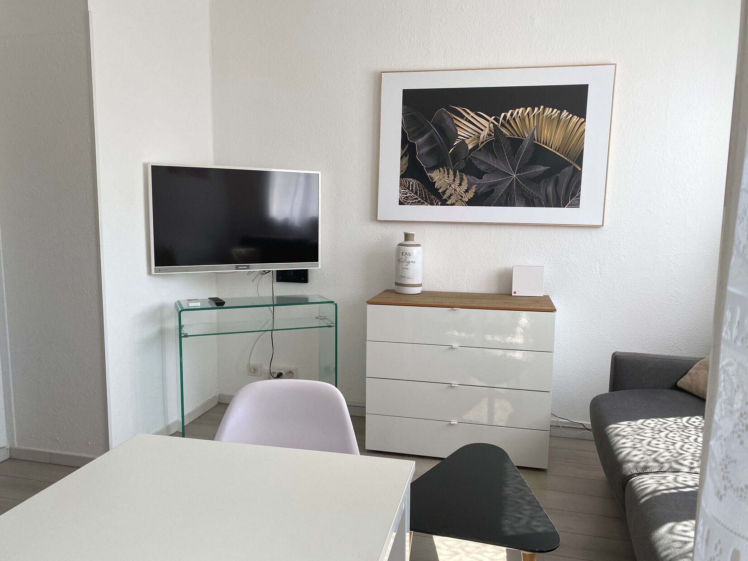 A modern living room with a flat-screen TV, a glass coffee table, a white sofa, a black armchair, a white dresser, and a framed botanical print on the wall.