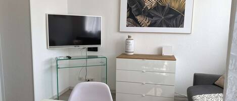 A modern living room with a flat-screen TV, a glass coffee table, a white sofa, a black armchair, a white dresser, and a framed botanical print on the wall.