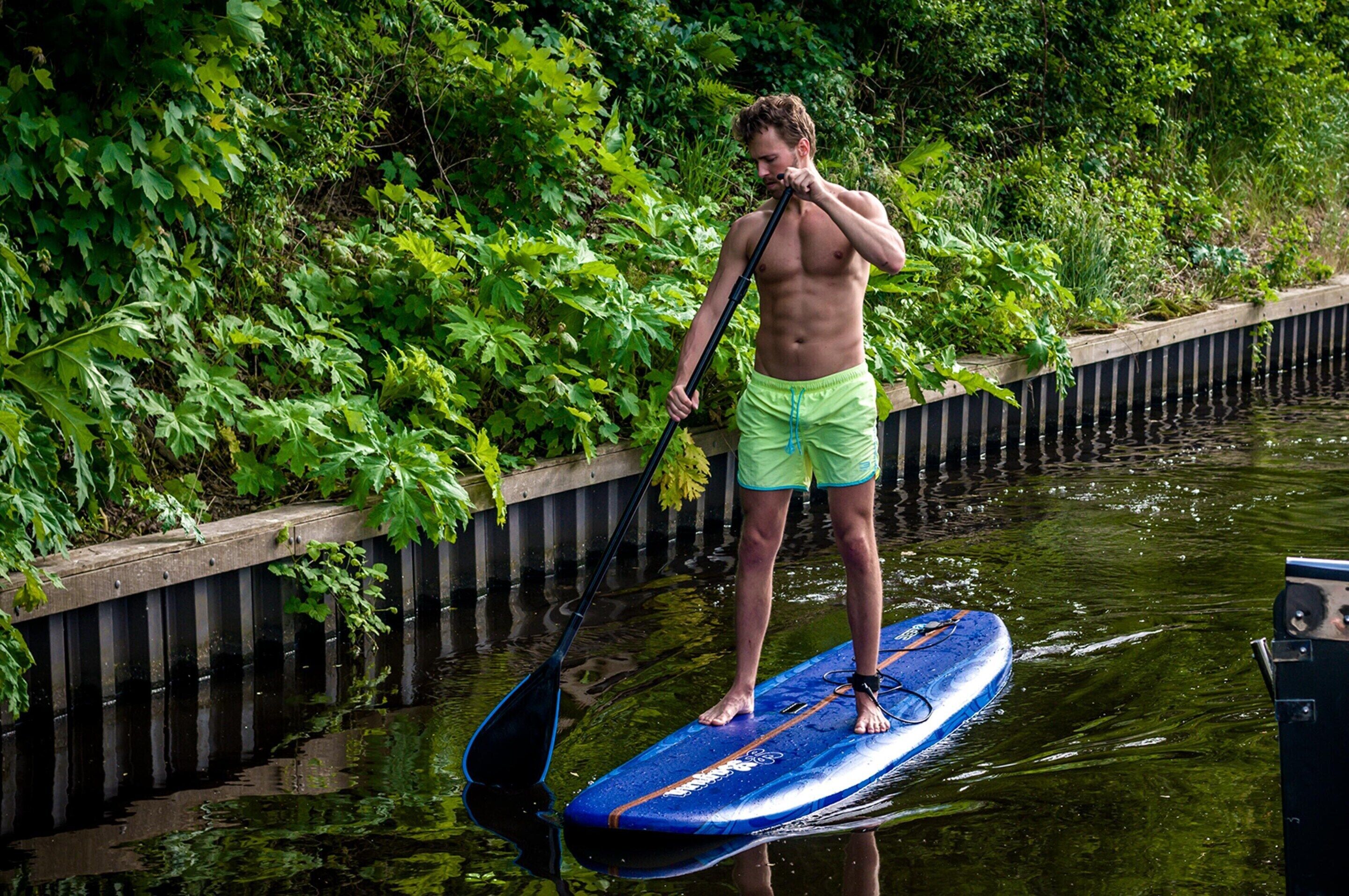 Eine Person steht auf einem Stand-Up-Paddleboard in einem von Grünpflanzen umgebenen Gewässer.
