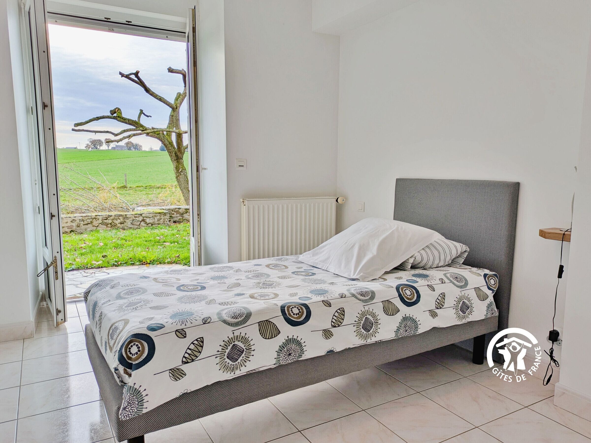 Un dormitorio con cama, un radiador y vistas a un árbol y un campo a través de una puerta abierta.