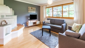 A modern living room with a flat-screen TV, grey sofa, wooden flooring, and a large window with a view of greenery. - Holiday Home in a Beautiful Location (Radstadt)