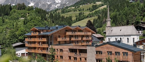 A wooden multi-story building with balconies, set against a backdrop of mountains and greenery.