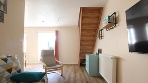 A living room with a patterned sofa, a wooden chair, a teal cabinet, and a TV mounted on the wall. - Semi-detached house with private spa, pets welcome, near Cotentin (Saussemesnil)