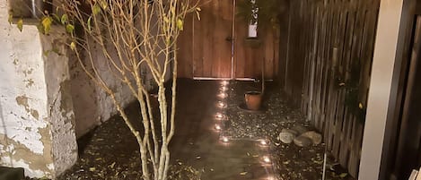 A narrow pathway with a tree on the left, a wooden fence on the right, and a door at the end.