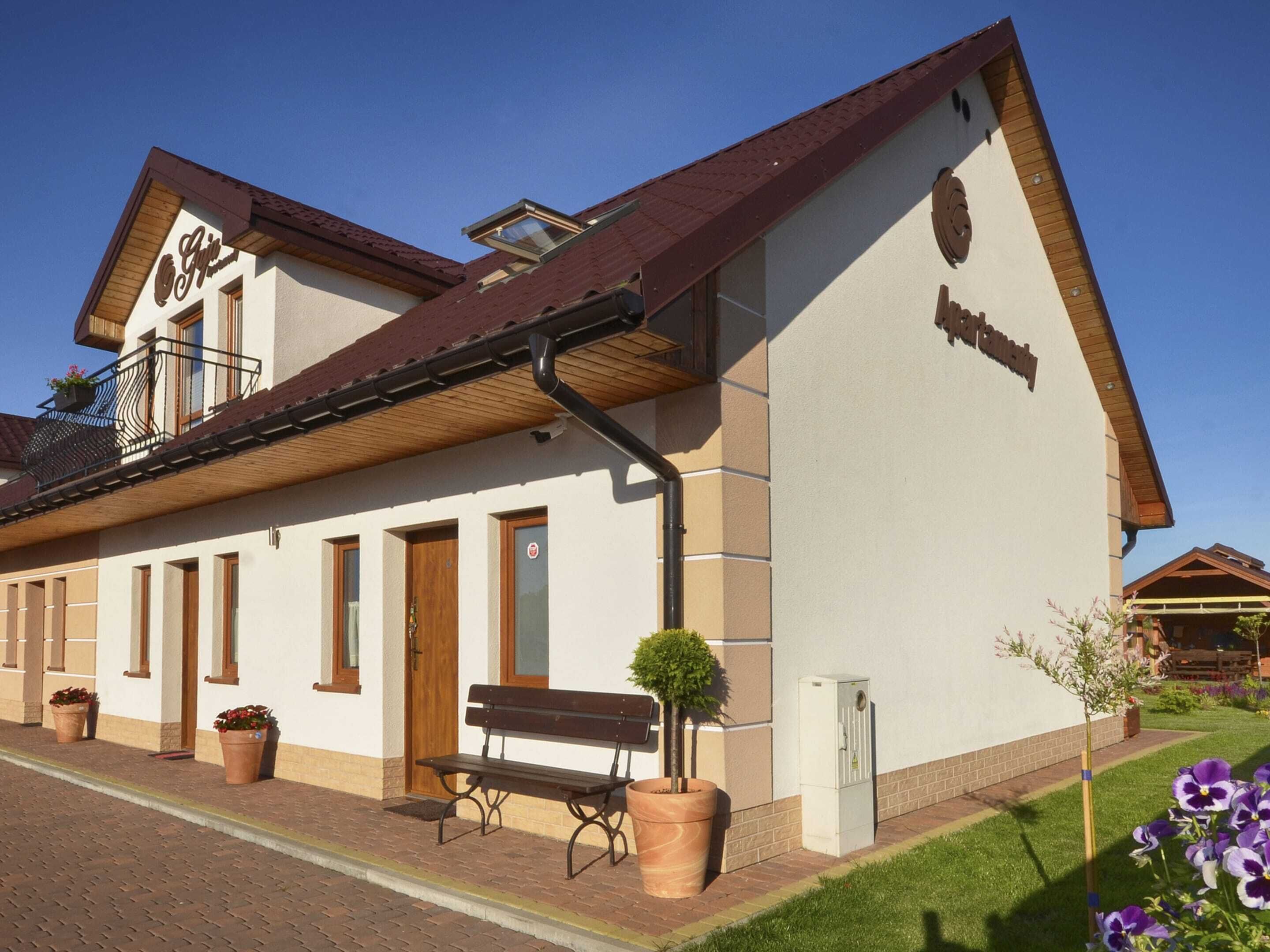 Un edificio con facciata bianca, tetto marrone e un balcone con ringhiera in legno. Davanti all'edificio, una panchina e piante in vaso.