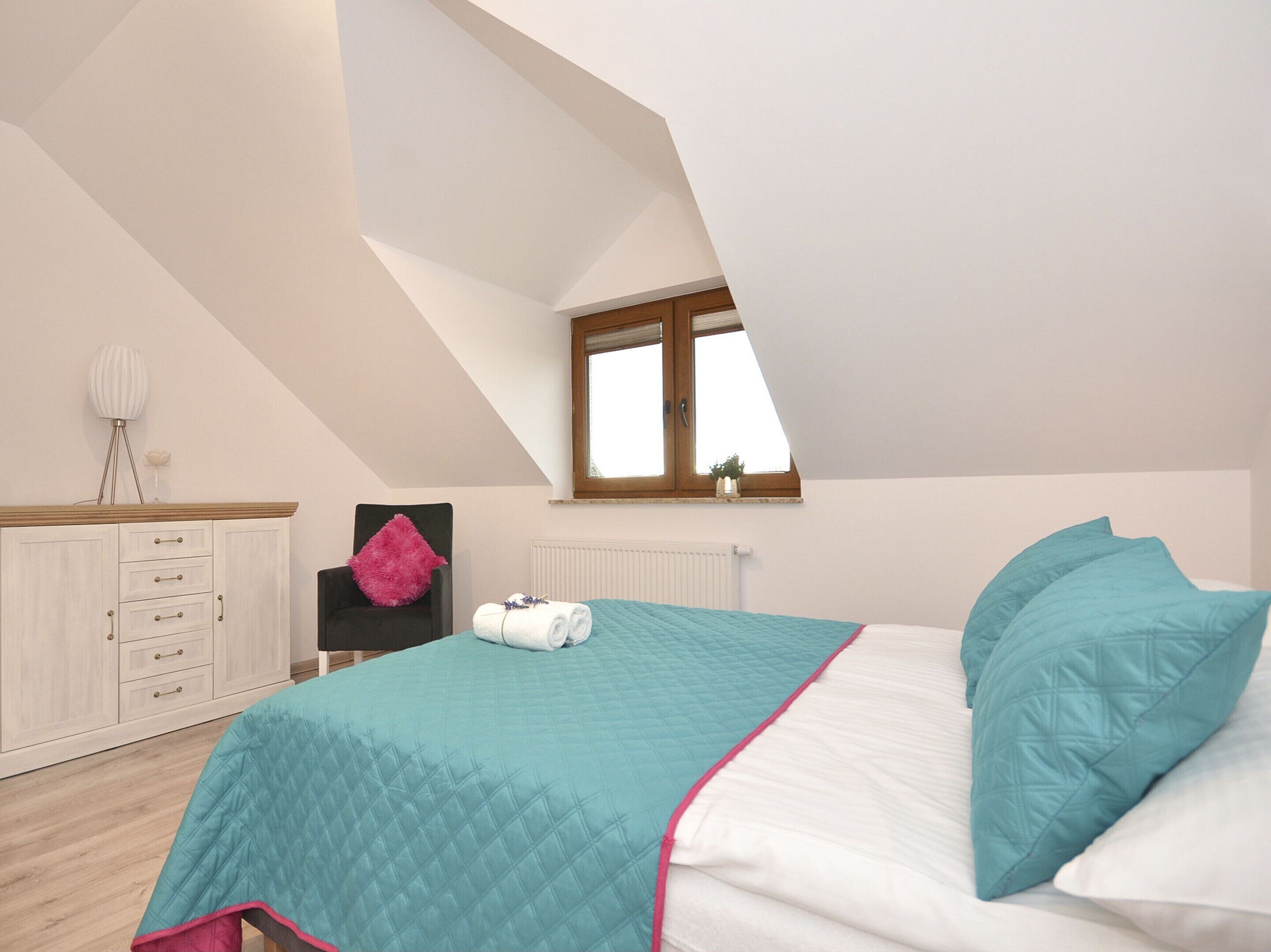 A bedroom with a sloped ceiling, a bed with teal and white bedding, a wooden dresser, a pink chair, and a window with a view.