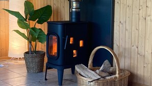 A room with a black wood-burning stove, a potted plant, and a wicker basket filled with logs. - Charming Stay by the Meuse (Hastiere)