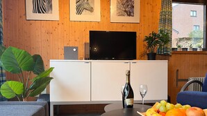 A modern living room with a wooden wall, a flat-screen TV, a white cabinet, a large plant, a blue chair, and a table with fruit and a bottle of champagne. - Charming Stay by the Meuse (Hastiere)