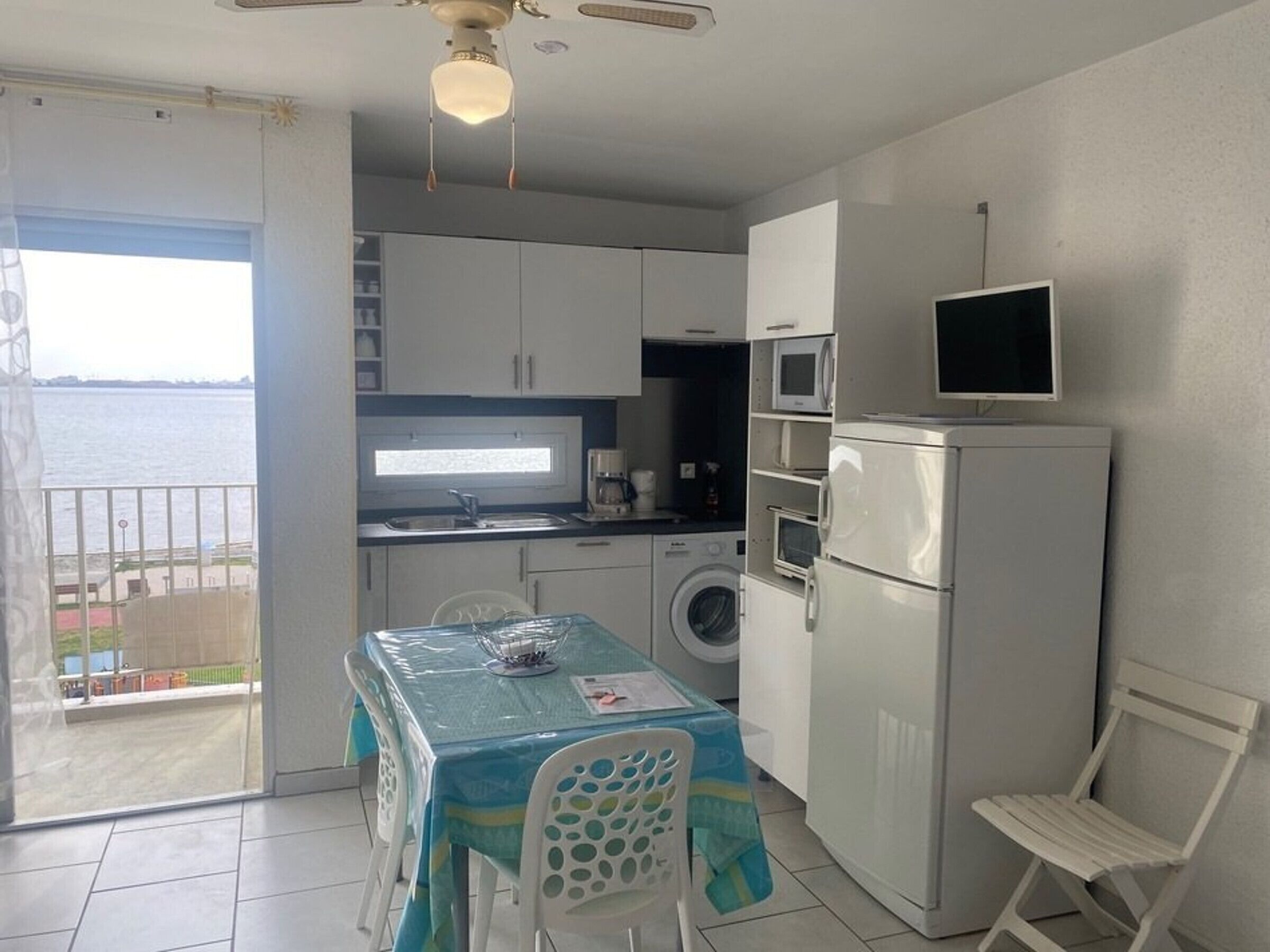 Une petite cuisine équipée avec un lave-linge, un four à micro-ondes et un réfrigérateur. On y trouve une table avec des chaises et une vue sur la mer, accessible par une porte coulissante.