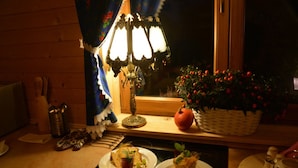 Une cuisine faiblement éclairée, avec des murs en bois, une table sur laquelle sont disposés des plats, une lampe et une fenêtre ornée de rideaux.