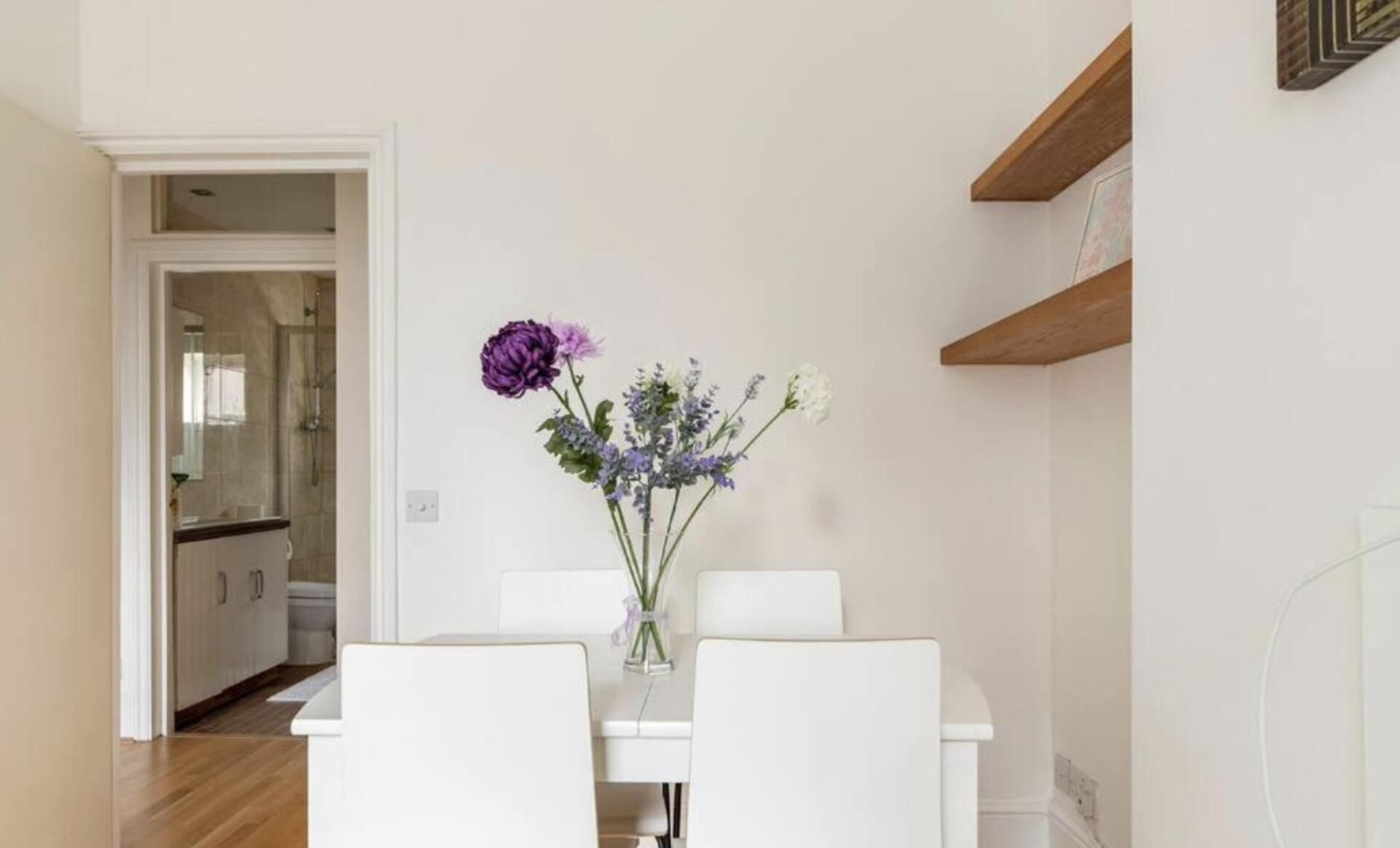 A dining area with a white table and chairs, a vase with flowers, and a kitchen in the background.