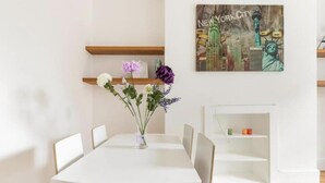 Ein Essbereich mit einem weißen Tisch, Stühlen und einer Blumenvase. An der Wand hängt ein Gemälde von New York City.