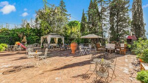 An outdoor patio with brick flooring, metal chairs, tables, a gazebo, and string lights.