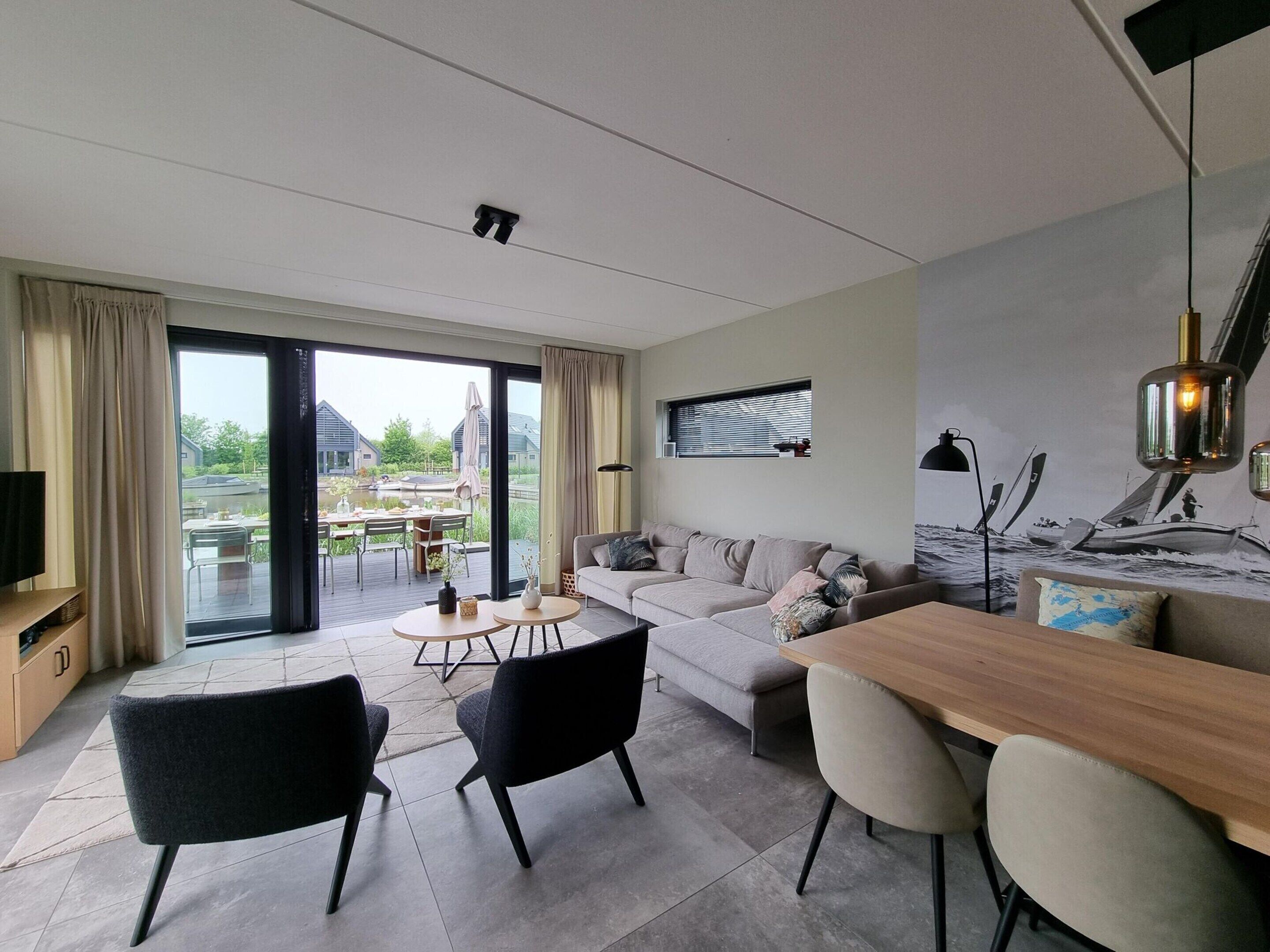 Un salon moderne comprenant un canapé, des fauteuils, une table à manger et offrant une vue sur un jardin et des bâtiments à l’extérieur.