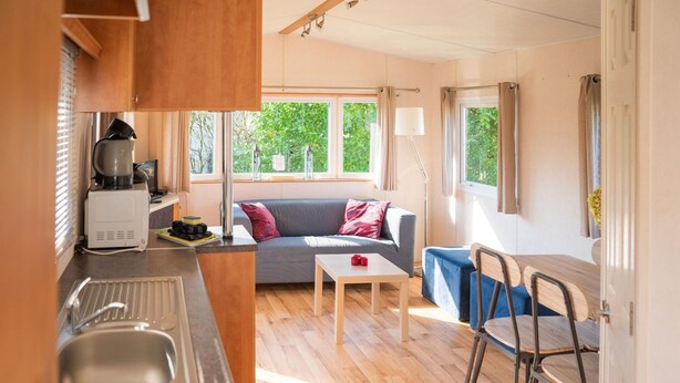 A compact kitchen with a stove, sink, and a coffee maker. There is a dining area with a table and chairs, and a sofa with cushions. Large windows provide natural light and a view of greenery outside. - Cosy Holiday Home in Appelscha With Garden (Appelscha)