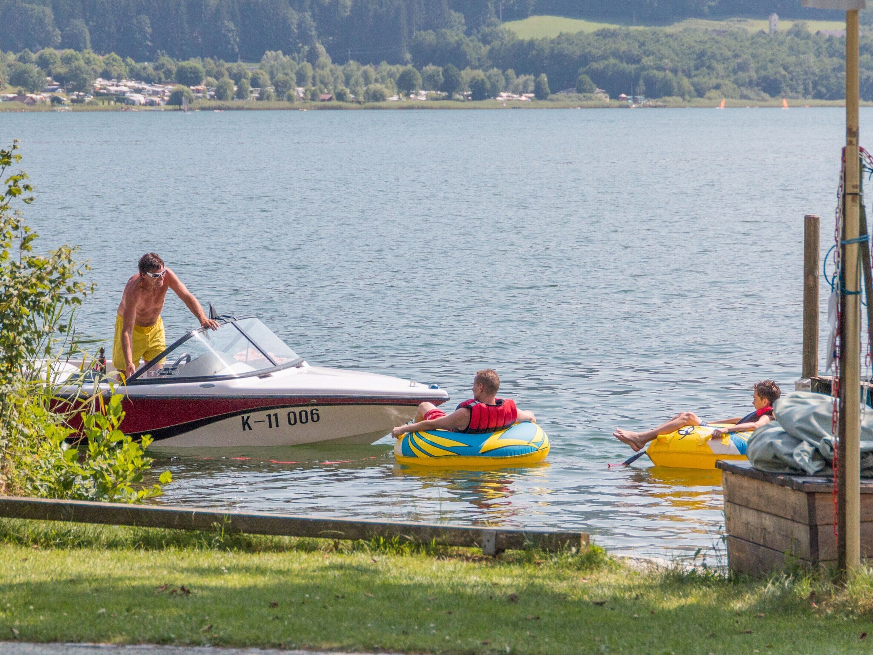 A lake with a boat docked at the shore, people on inflatable rafts, and a grassy area in the foreground.