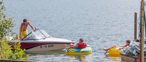 A lake with a boat docked at the shore, people on inflatable rafts, and a grassy area in the foreground.