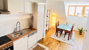 Een kleine keuken met een spoelbak, fornuis en kastjes, die uitkomt op een eethoek met een tafel en stoelen.