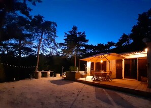Uma área de pátio externa com uma mesa e cadeiras de jantar, cercada por árvores e um guarda-sol bem iluminado.