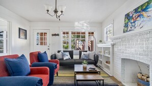 Ein Wohnzimmer mit einer Couch, einem roten Sessel und einem Couchtisch. Ein Kamin mit weißem Ziegelumfang und ein Bild darüber sind ebenfalls zu sehen.
