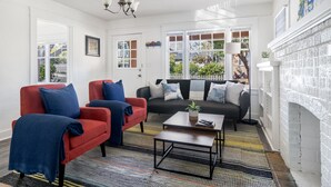 Ein Wohnzimmer mit einem roten Sessel, einem schwarzen Sofa und einem Couchtisch. Rechts befindet sich ein Kamin und eine Tür zum Garten.