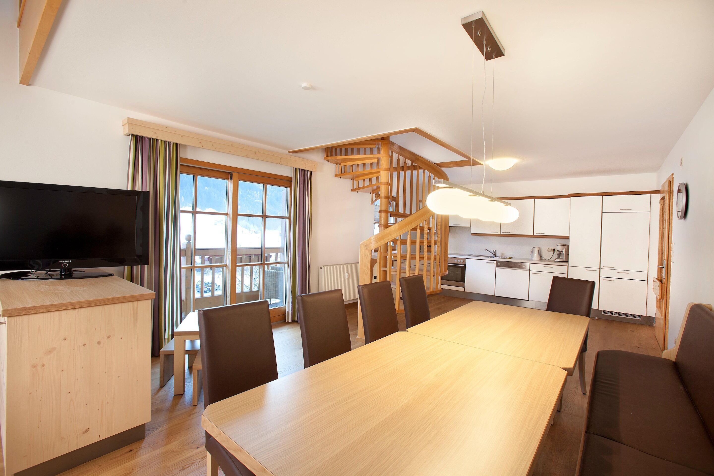 A modern living space with a wooden dining table, chairs, a TV, a kitchen area, and a staircase.