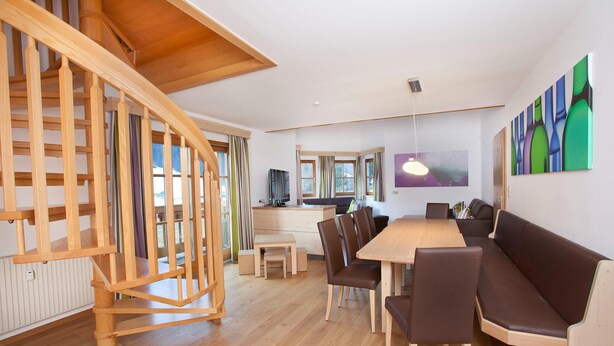 A modern interior with a wooden staircase, a dining area with a long table and chairs, and a seating area with a sofa.