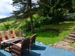 An outdoor seating area with a wooden table and chairs, a checkered cushion, and a view of a lush green landscape with trees and a hill in the background. - Stylish Retreat in Harz (Herzberg am Harz)