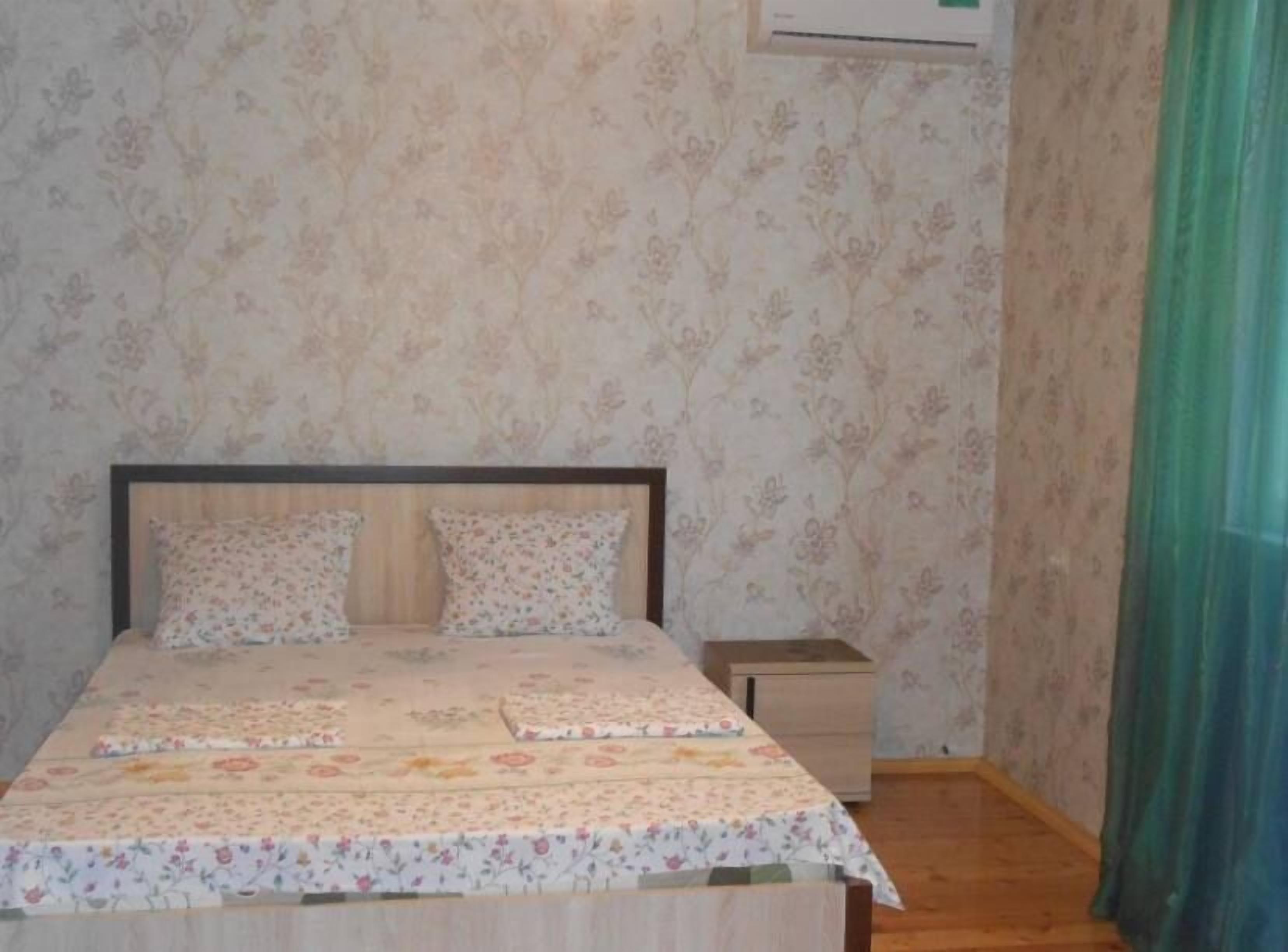 A bedroom with floral wallpaper, a bed with a floral bedspread, a nightstand, and a small drawer unit.