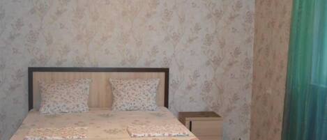 A bedroom with floral wallpaper, a bed with a floral bedspread, a nightstand, and a small drawer unit.