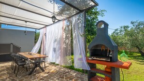 Een terras buiten met een stenen vloer, een tafel met stoelen, een barbecue en een overdekt gedeelte met dunne gordijnen.