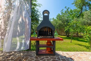 An outdoor stone oven with a chimney, placed on a red platform with a stone base, surrounded by a grassy area and trees. - Holiday house Chill out (Marcana)