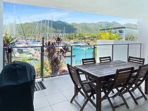 A balcony with a table and chairs, a covered chair, and a view of a marina with boats and mountains.