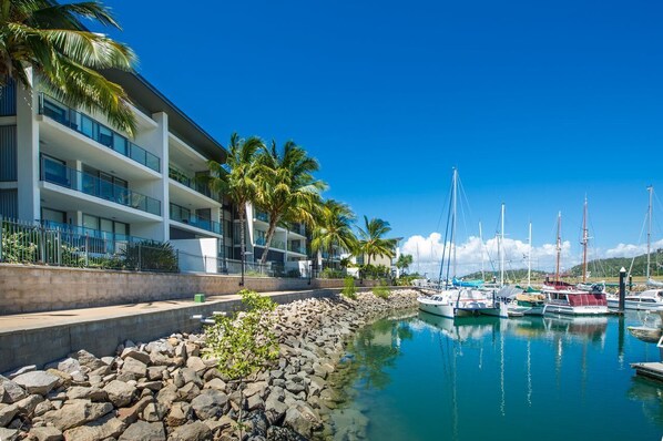 A marina with boats, a modern building with balconies, and palm trees.