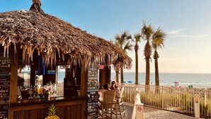 Un bar en la playa con techo de paja, mostrador de bar de madera y una pizarra con el menú.