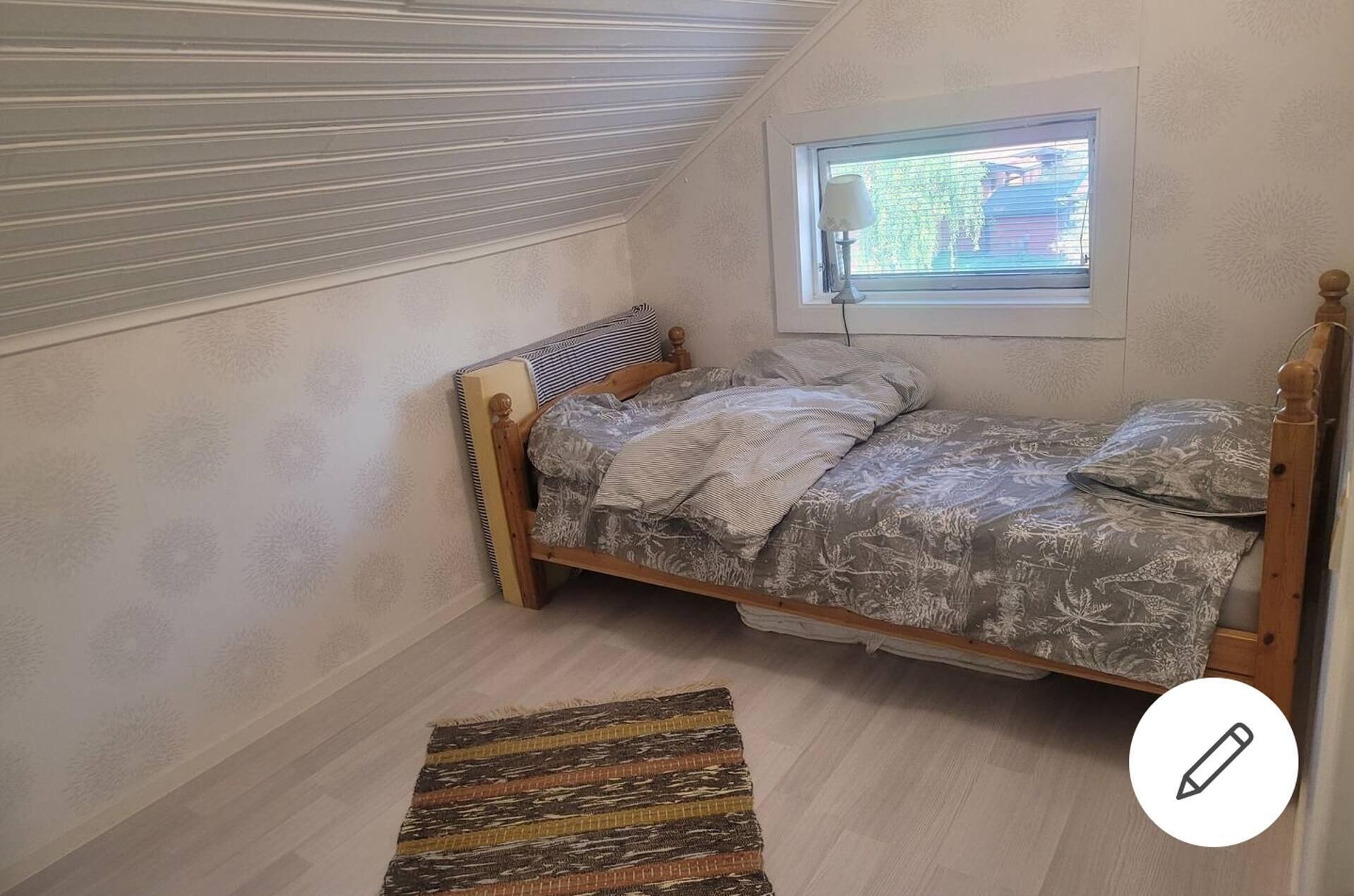 A small, single-bed room with a sloped ceiling, a window with blinds, and a patterned rug on the floor.