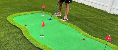 A person playing mini-golf in a backyard with a green artificial turf course, white fence, and a house in the background.