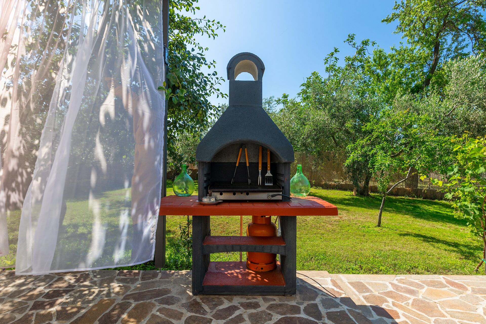A stone-built outdoor pizza oven with a chimney, a table with a bottle, and a grill on a stone patio surrounded by greenery.