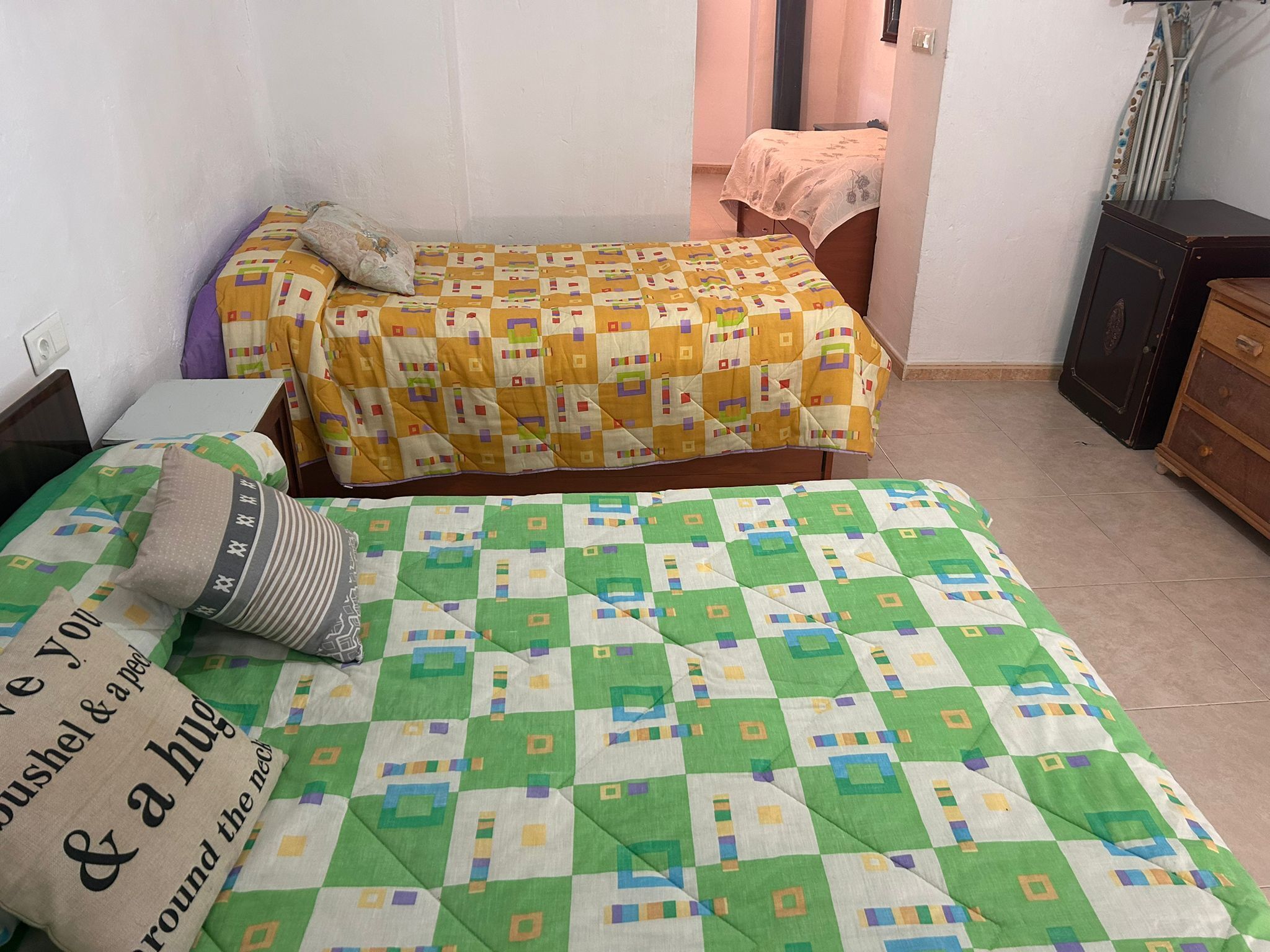 Une chambre avec deux lits : l’un est recouvert d’une couvre-lit à carreaux jaunes et blancs, et l’autre d’une couverture à motifs géométriques verts et blancs. On y trouve une commode en bois et une table de chevet avec une lampe.