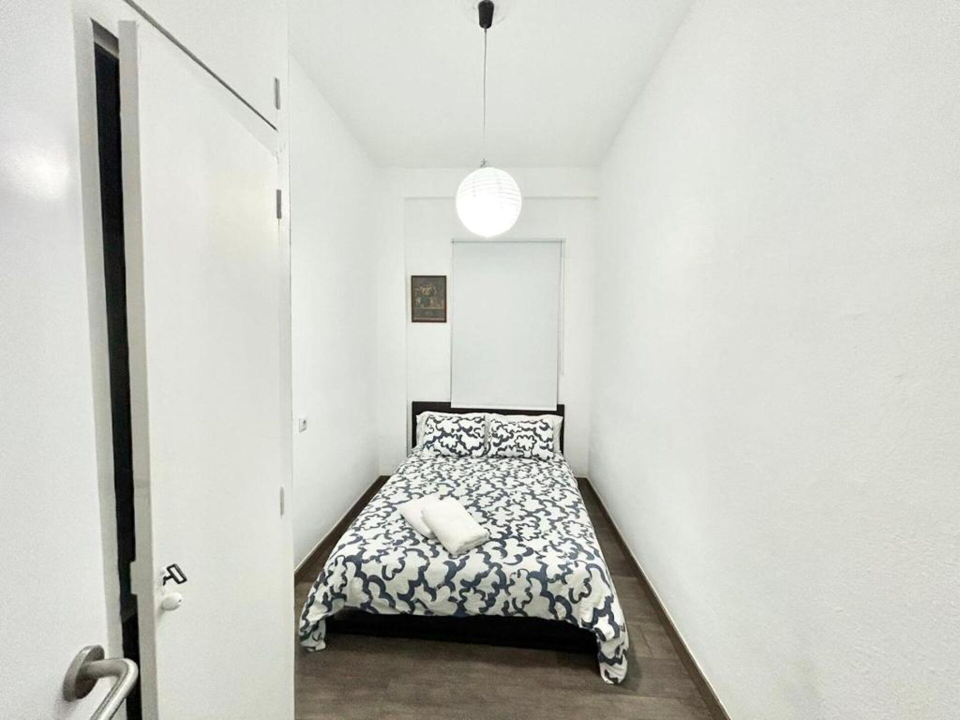 Un couloir étroit mène à un lit recouvert d’une couvre-lit à motifs, un luminaire rond est suspendu au plafond, et une porte dotée d’une poignée se trouve à la fin du couloir.