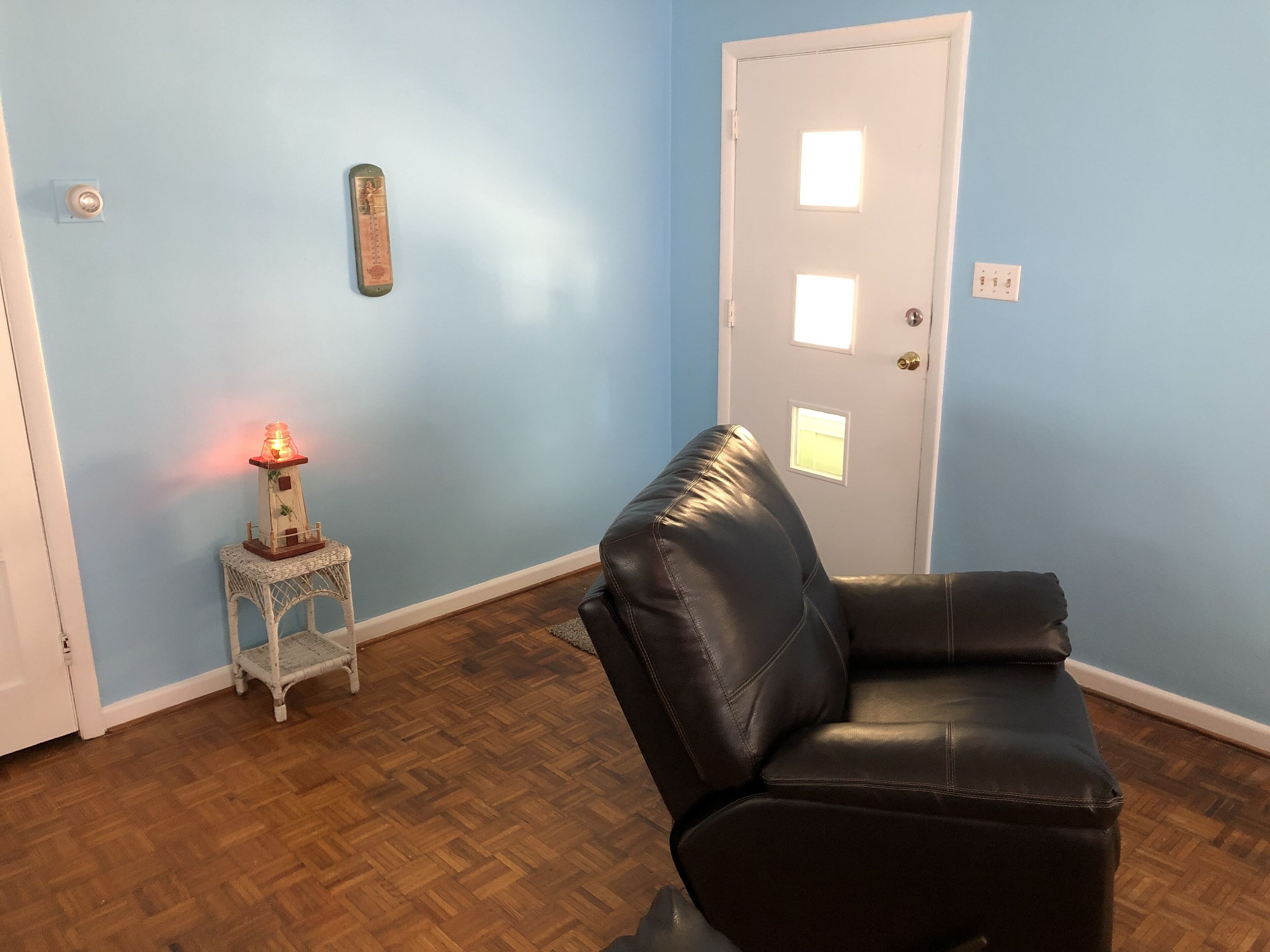 Une pièce avec un plancher en bois, un fauteuil en cuir noir, une petite table blanche ornée d’un objet décoratif et une porte blanche avec une fenêtre.