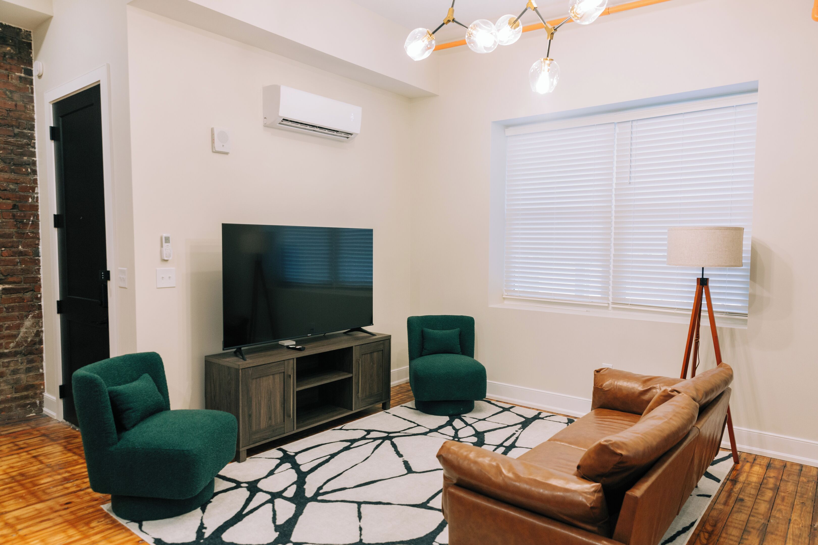 A modern living room with a flat-screen TV, two green armchairs, a brown leather sofa, a patterned rug, and a tripod lamp.