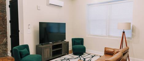 A modern living room with a flat-screen TV, two green armchairs, a brown leather sofa, a patterned rug, and a tripod lamp.