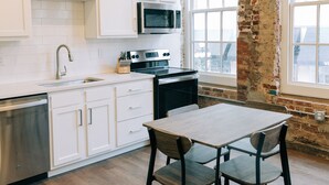 A modern kitchen with white cabinets, a stainless steel dishwasher, a microwave, and a brick wall. There is a dining table with four chairs. - The St. Clair (Frankfort)