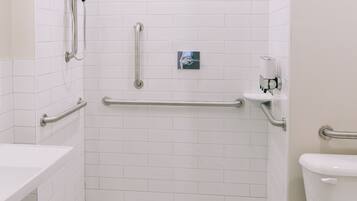 Un baño con paredes de azulejos blancos, inodoro blanco, lavamanos y zona de ducha con barras de apoyo.