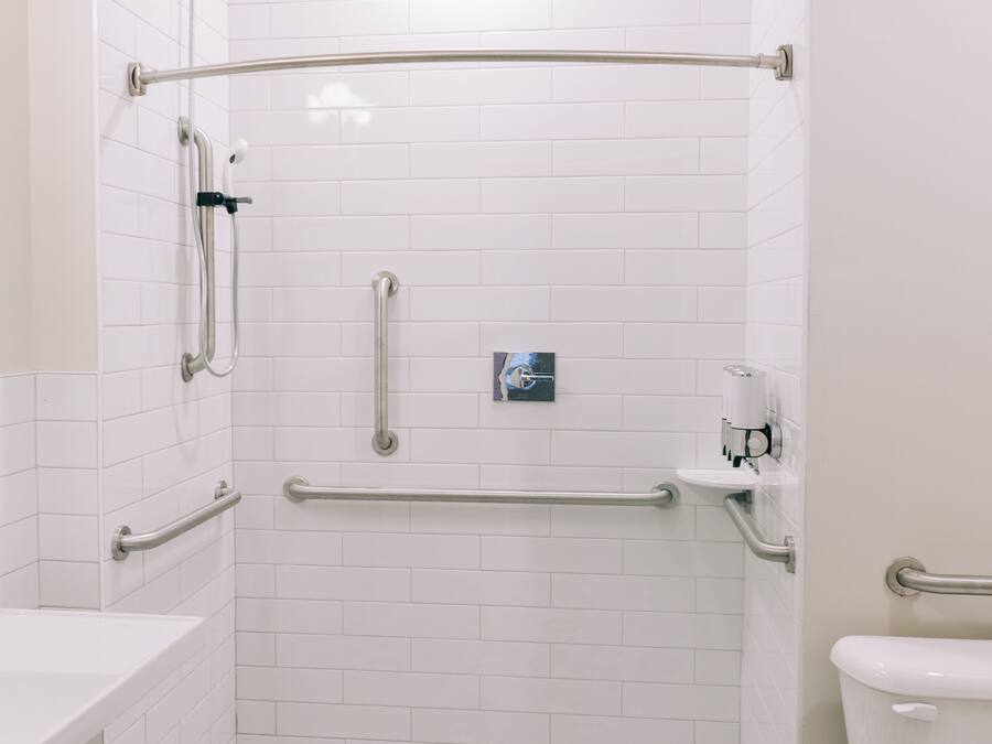 Un baño con paredes de azulejos blancos, inodoro blanco, lavamanos y zona de ducha con barras de apoyo.