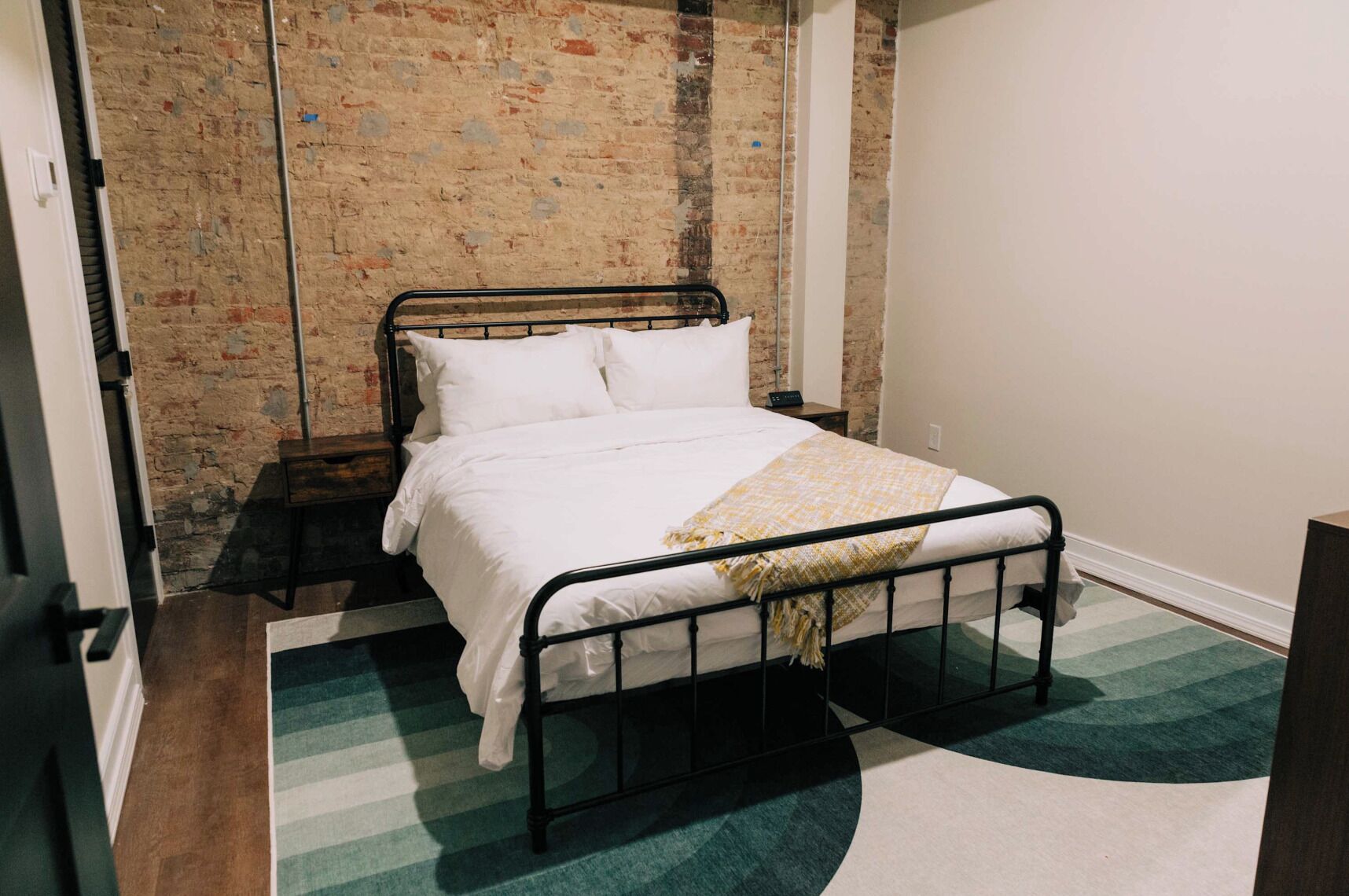 Een enkel bed met een metalen frame, wit beddengoed en een deken in geel en wit. De kamer heeft een bakstenen muur en een vloerkleed met groene en witte strepen.