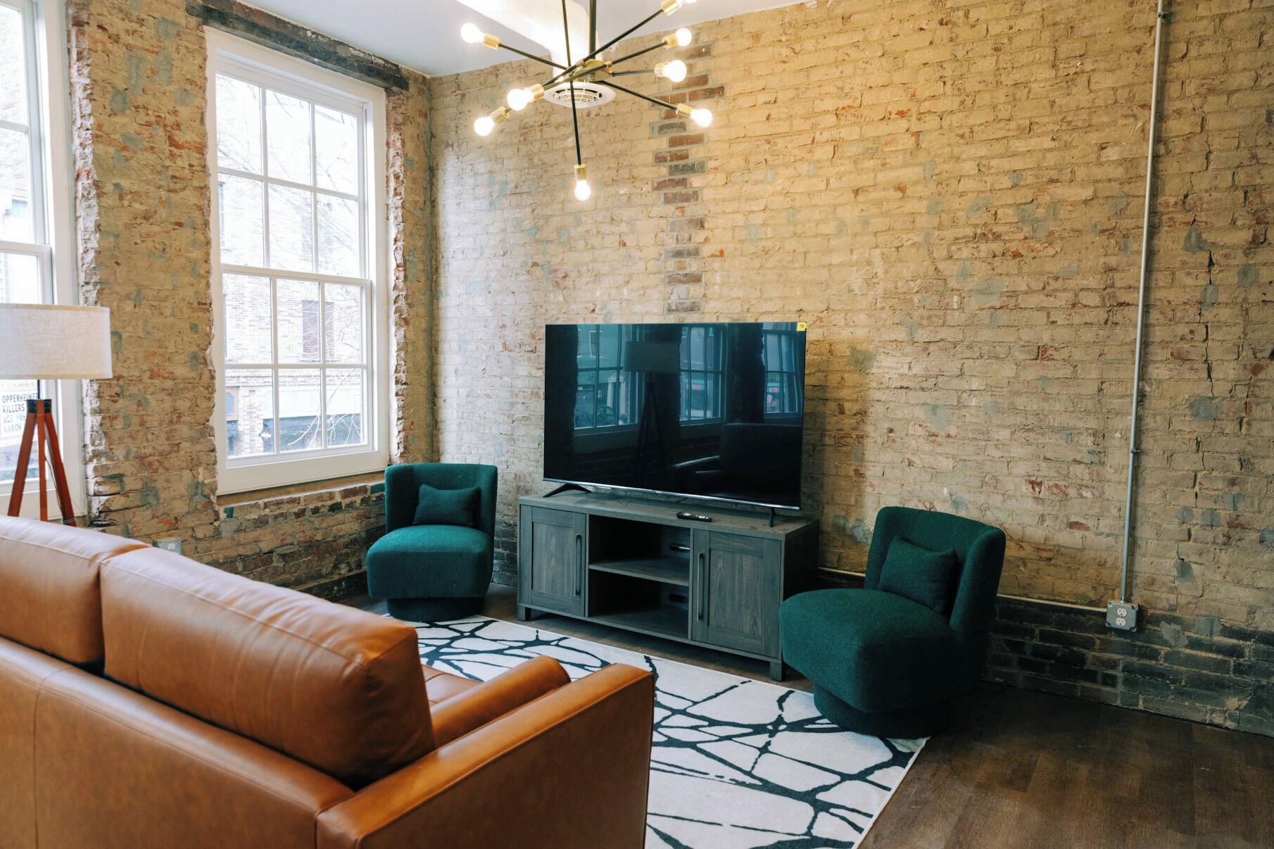 A living room with a brick wall, a large flat-screen TV, two green armchairs, a brown leather sofa, and a modern chandelier.