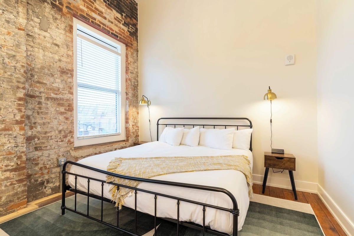 Un dormitorio con pared de ladrillo, un ventanal con persianas, estructura de cama metálica, ropa de cama blanca, una lámpara de noche y una mesita de noche de madera.